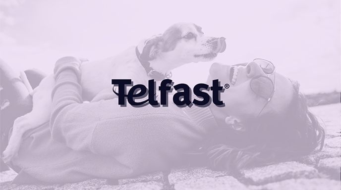 A promotional image with a soft-toned filter, featuring the Telfast® logo over a background of a joyful woman in a sweater and sunglasses lying on the ground, laughing as a small dog playfully licks its nose above her.