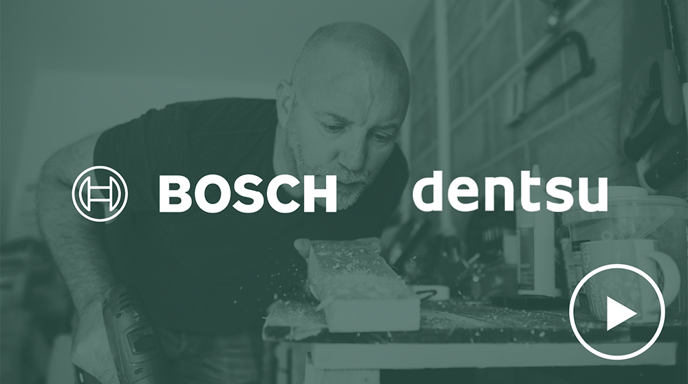Ein Mann mit Bauhelm, der ein blaues T-Shirt trägt, benutzt eine Bohrmaschine von Bosch auf einer Baustelle. Die Logos von Bosch und Dentsu befinden sich in der oberen linken Ecke