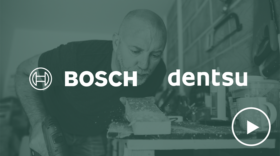 Ein Mann mit Bauhelm, der ein blaues T-Shirt trägt, benutzt eine Bohrmaschine von Bosch auf einer Baustelle. Die Logos von Bosch und Dentsu befinden sich in der oberen linken Ecke